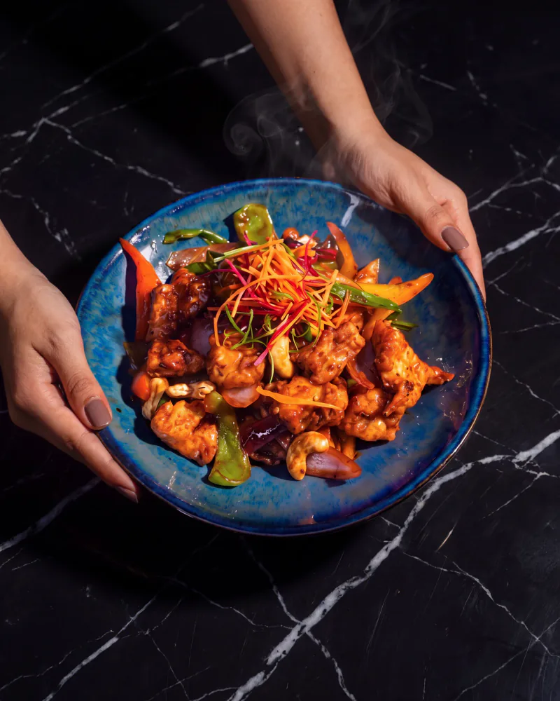 Wok-fried chicken with cashews and peppers in a blue glazed bowl, held over black marble