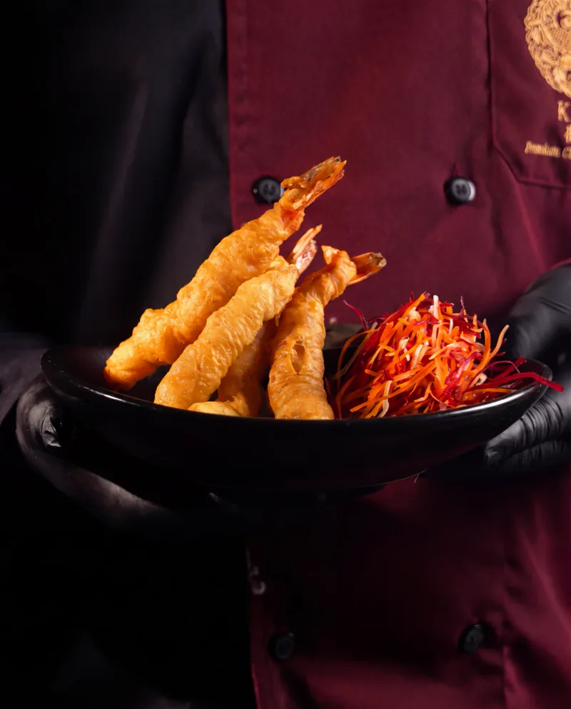 A KAO chef in a maroon jacket and black gloves holding a bowl of prawn tempura